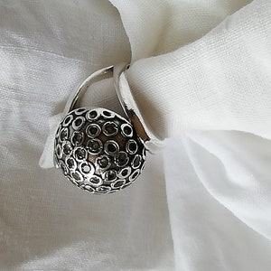 May include: A silver ring with a round, textured top. The top of the ring is covered in small, black circles.