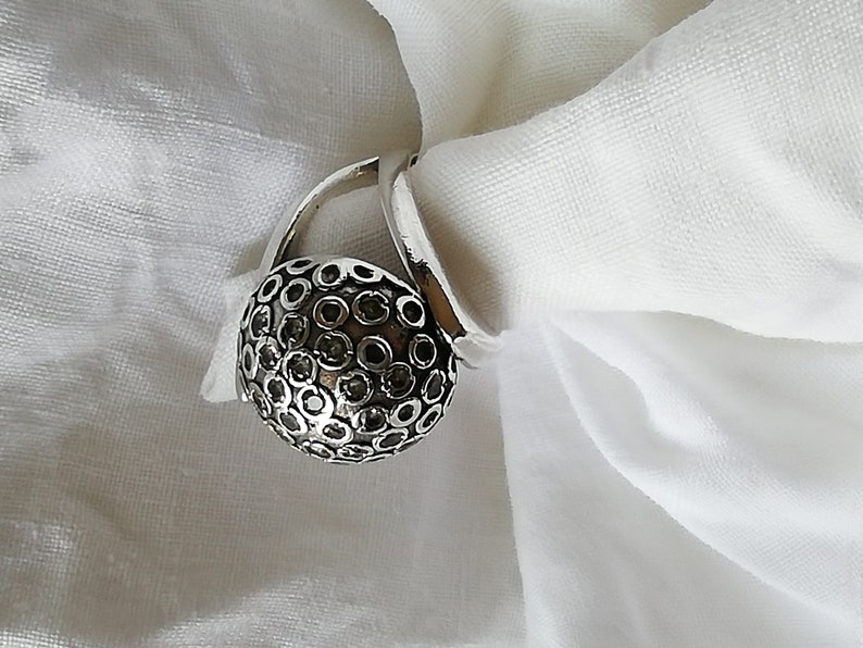May include: A silver ring with a round, textured top. The top of the ring is covered in small, black circles.
