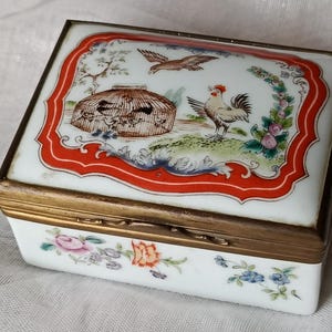 May include: Rectangular white trinket box with a hinged lid and brass trim. The lid features a painted scene with a rooster, birdcage, and flying bird, framed by a red border. Floral designs adorn the box's sides.