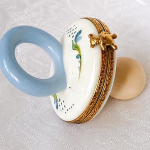 May include: A porcelain pacifier-shaped trinket box with a glossy white finish, featuring a light blue handle, hand-painted green and blue floral accents, a gold-toned metal hinge, and a small gold-toned teddy bear figurine attached to the rim.