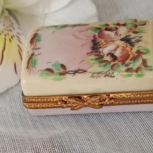 May include: A rectangular trinket box with a cream-colored lid and a pink base. The lid features a floral design with pink and brown roses and green leaves. The box has gold-colored metal trim and a decorative clasp.