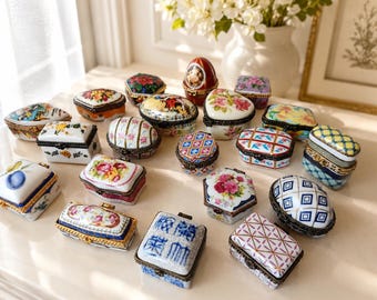 May include: A collection of small, hinged porcelain trinket boxes in various shapes, including oval, rectangular, and heart-like forms. The boxes feature diverse patterns such as floral motifs, geometric grids, and blue and white designs.
