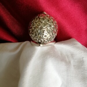 May include: A silver ring with a round, textured top. The top of the ring is covered in small, circular designs.