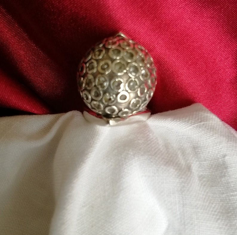 May include: A silver ring with a round, textured top. The top of the ring is covered in small, circular designs.