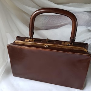 May include: A vintage brown leather handbag with a curved wooden handle. The rectangular bag features a gold-toned clasp and trim. The bag is set against a white background.