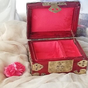 May include: A red wooden jewelry box with brass hardware, open to reveal a bright pink interior. The box has a rectangular shape with decorative corner accents and a clasp. A pink flower rests nearby.