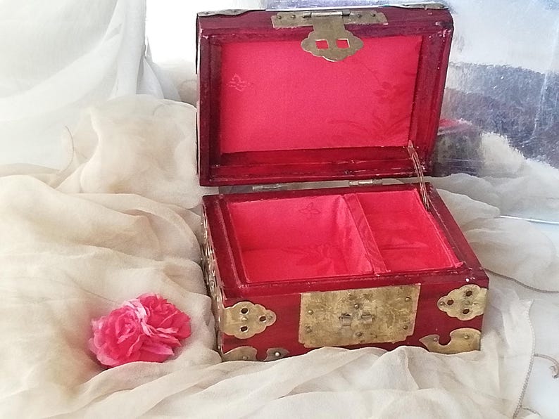 May include: A red wooden jewelry box with brass hardware, open to reveal a bright pink interior. The box has a rectangular shape with decorative corner accents and a clasp. A pink flower rests nearby.