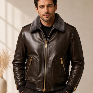 May include: A dark brown leather bomber jacket featuring a grey shearling collar, a central gold-tone zipper, two zippered front pockets, and ribbed cuffs. The jacket includes gold-tone zipper details on the sleeves and a black ribbed hem.