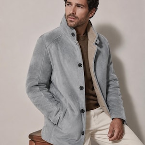 May include: A light gray suede jacket with a high collar, dark buttons, and a beige shearling lining. The jacket is worn over a brown ribbed sweater and paired with off-white trousers, creating a layered look with visible texture and stitching.