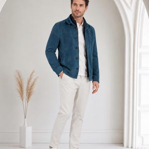 May include: A teal blue suede jacket with a pointed collar and dark buttons, worn over a white polo shirt. The outfit includes light beige trousers and white sneakers. The jacket features long sleeves and a structured fit, paired with neutral pants.