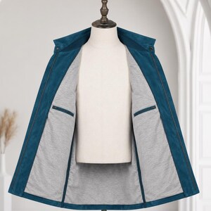 May include: A teal jacket displayed on a mannequin, featuring a fold-down collar and a full-length front zipper. The interior is lined with a light gray fabric and includes two horizontal zippered pockets on the inner chest panels.