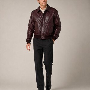 May include: A dark burgundy leather bomber jacket with a collar, shoulder epaulets, and front flap pockets, worn over a white button-down shirt, paired with dark gray trousers and black leather dress shoes.