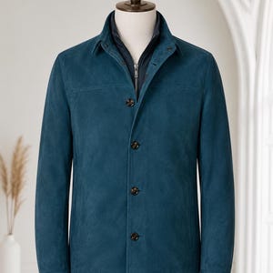 May include: A teal suede jacket featuring a structured collar, long sleeves, and a front button closure. The jacket is layered over a dark navy blue zip-up inner piece, creating a dual-layer appearance on a mannequin torso.