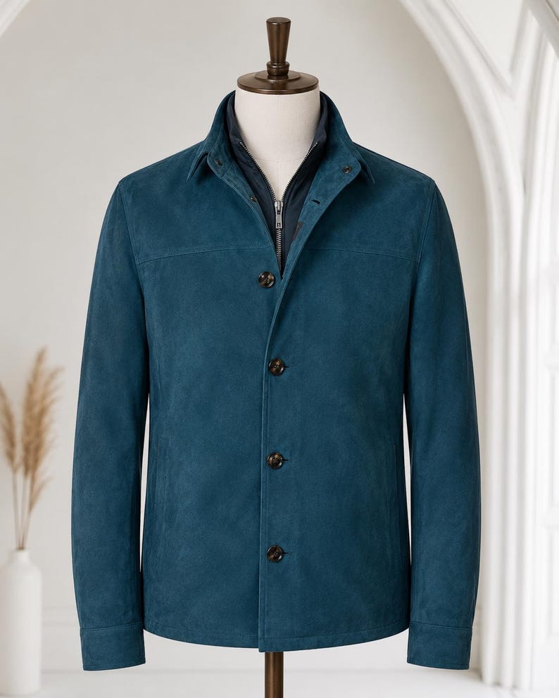 May include: A teal suede jacket featuring a structured collar, long sleeves, and a front button closure. The jacket is layered over a dark navy blue zip-up inner piece, creating a dual-layer appearance on a mannequin torso.