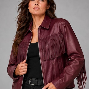 May include: A deep burgundy leather jacket featuring a classic collar, a full-length silver-tone front zipper, and long fringe detailing across the chest and down the outer sleeves. The jacket is worn over a simple black top and black jeans.