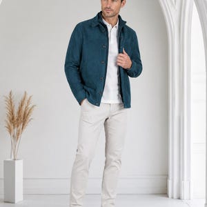 May include: A teal suede jacket with a button-down front and collar, worn over a white polo shirt. The outfit is completed with light beige trousers and white sneakers, creating a coordinated look with clean lines and a textured fabric finish.
