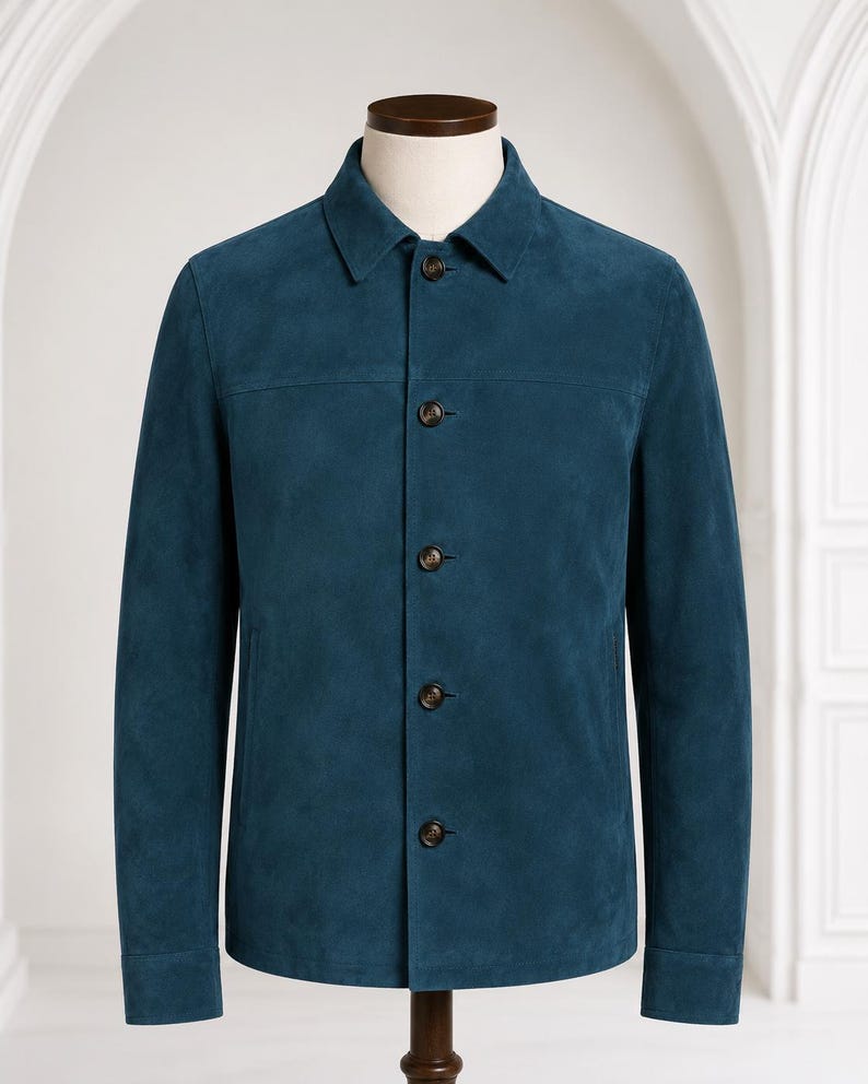 May include: A teal blue suede jacket featuring a classic point collar, long sleeves with buttoned cuffs, and a front button placket with five dark brown buttons. The garment has a horizontal seam across the chest and two side pockets.