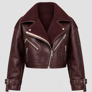 May include: A cropped burgundy leather jacket featuring a wide lapel collar lined with dark maroon faux shearling. The jacket includes silver-tone metal zippers on the front and pockets, along with adjustable buckled straps at the sleeve cuffs.