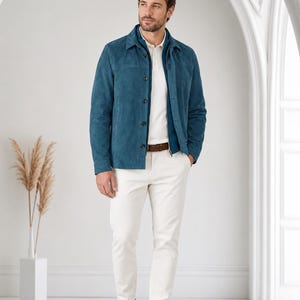 May include: A teal suede jacket with a pointed collar and dark buttons, worn over a white polo shirt and white trousers. The outfit includes a brown leather belt and brown suede sneakers, creating a coordinated look with clean lines and textures.