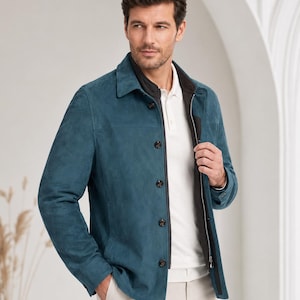 May include: A teal suede jacket featuring a classic collar, long sleeves, and a front button closure. The jacket is layered over a white polo shirt and light-colored trousers. The design includes a dark inner lining and visible stitching details.