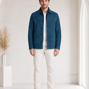 May include: A teal suede jacket with a pointed collar and dark buttons, worn over a white collared shirt and a dark zip-up layer. The outfit is completed with off-white trousers and white sneakers, creating a layered look with a textured finish.