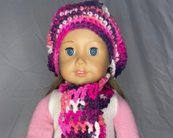 May include: A doll wearing a pink and purple crocheted hat and scarf. The hat is a beret style and the scarf has long fringe. The doll is wearing a pink sweater and a white shirt.