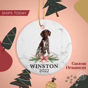 May include: A white ceramic custom ornament with a marble design, featuring a brown and white dog illustration. The ornament includes the name "WINSTON" and the year "2022". A pink bow and greenery are at the bottom. The text "Custom Ornament" is on the right.