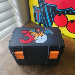 May include: A rectangular black storage case with two bright orange plastic latches on the front. The lid features a graphic of a red number 3, a white and gray circular object, and a cartoon orange dragon character with blue wings and a flame tail.