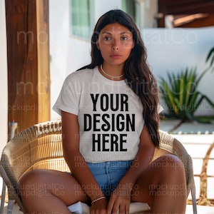 May include: A woman wearing a white t-shirt with the text "YOUR DESIGN HERE" printed on it. She is sitting on a wicker chair with her legs crossed.