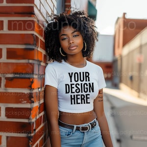 May include: A woman wearing a white crop top with the text "YOUR DESIGN HERE" printed on it. She is standing next to a brick wall and wearing blue jeans and a brown belt.