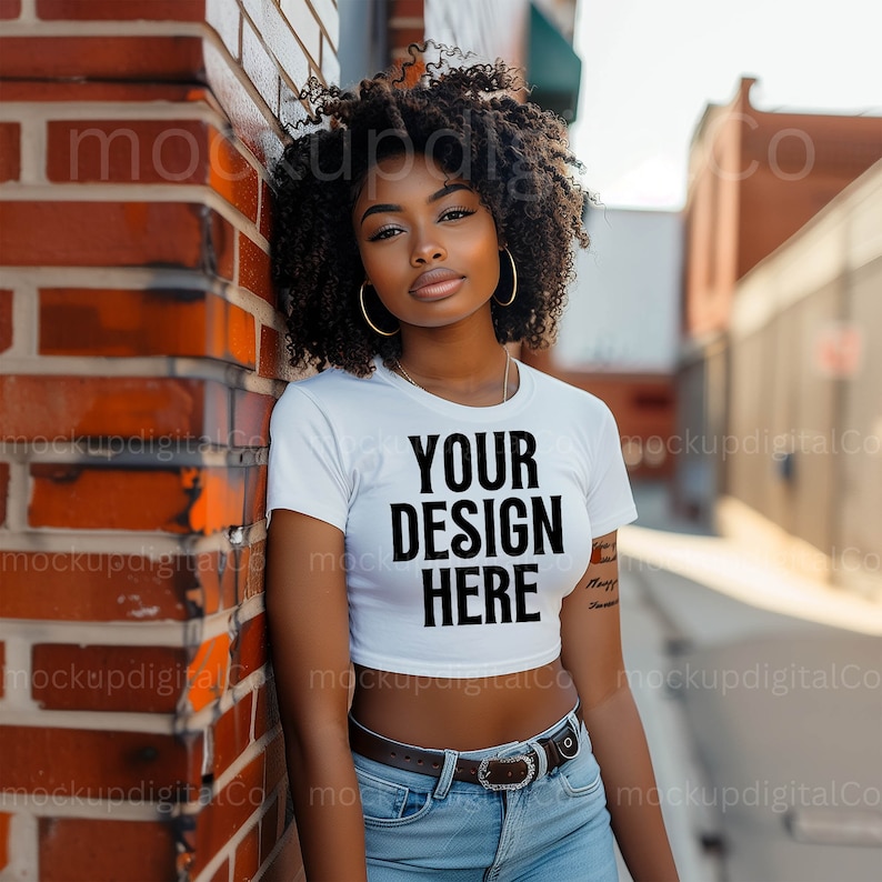 May include: A woman wearing a white crop top with the text "YOUR DESIGN HERE" printed on it. She is standing next to a brick wall and wearing blue jeans and a brown belt.