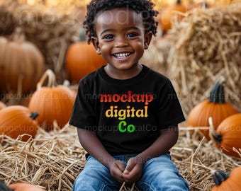 Könnte beinhalten: Ein lächelndes Kind trägt ein schwarzes T-Shirt mit den Worten "mockup digital Co" in Rot, Gelb und Grün. Das Kind sitzt auf Heu mit Kürbissen im Hintergrund. Das Kind trägt eine blaue Jeans.