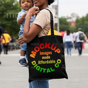 May include: Black tote bag with the text "MOCKUP Images made Affordable DIGITAL Co" in a circular design. The bag is carried by a person holding a child. The background shows a blurred outdoor scene with people.