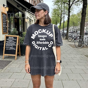 May include: A dark gray t-shirt with the text "MOCKUP Images made Affordable DIGITAL CO" in a white circular design. The person is wearing a gray baseball cap and carrying a white tote bag. The background shows a city street with trees and parked bicycles.