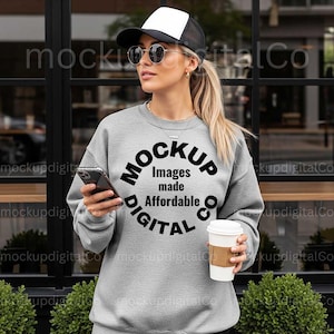 May include: A woman wearing a gray sweatshirt, black and white trucker hat, and sunglasses. The sweatshirt has a circular graphic that reads "MOCKUP Images made Affordable DIGITAL CO." She holds a smartphone and a coffee cup.