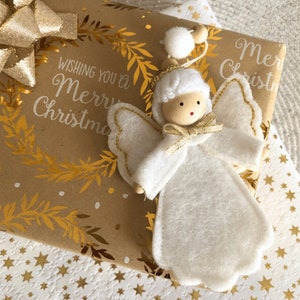 Può includere: Un ornamento di angelo in feltro bianco con finiture dorate e una testa di legno. L'angelo è seduto su carta da regalo marrone con accenti dorati in foglia e il testo "Wishing you a Merry Christmas".