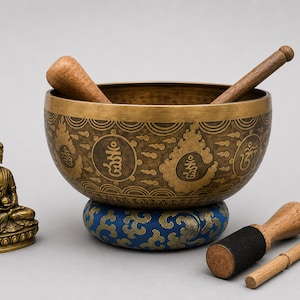 May include: A brass singing bowl with etched Tibetan script and cloud motifs, resting on a blue and gold fabric cushion. A small bronze Buddha statue sits to the left, accompanied by two wooden strikers, one featuring a black felt tip.