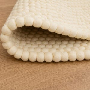 K&ouml;nnte beinhalten: Ein cremefarbener Teppich, der aus zahlreichen kleinen, kugelf&ouml;rmigen Filzwollkugeln in einem dichten, gleichm&auml;&szlig;igen Rastermuster besteht. Der Teppich ist teilweise umgeschlagen und zeigt die konsistente Textur und runde Form der Filzkugeln.