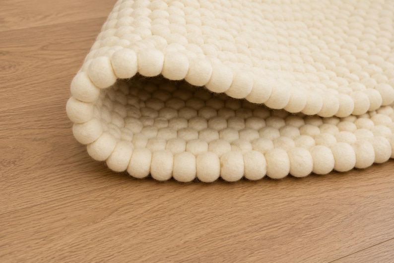 K&ouml;nnte beinhalten: Ein cremefarbener Teppich, der aus zahlreichen kleinen, kugelf&ouml;rmigen Filzwollkugeln in einem dichten, gleichm&auml;&szlig;igen Rastermuster besteht. Der Teppich ist teilweise umgeschlagen und zeigt die konsistente Textur und runde Form der Filzkugeln.