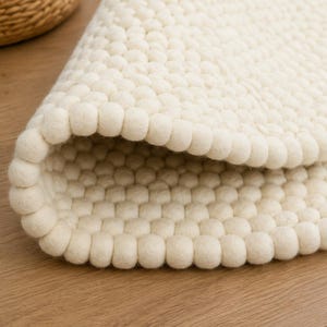 K&ouml;nnte beinhalten: Ein cremefarbener Teppich, der aus zahlreichen kleinen, kugelf&ouml;rmigen Filzwollkugeln in einem dichten, sich wiederholenden Muster besteht. Der Teppich ist teilweise umgeschlagen und zeigt die gleichm&auml;&szlig;ige Textur der Filzkugeln auf beiden Seiten.