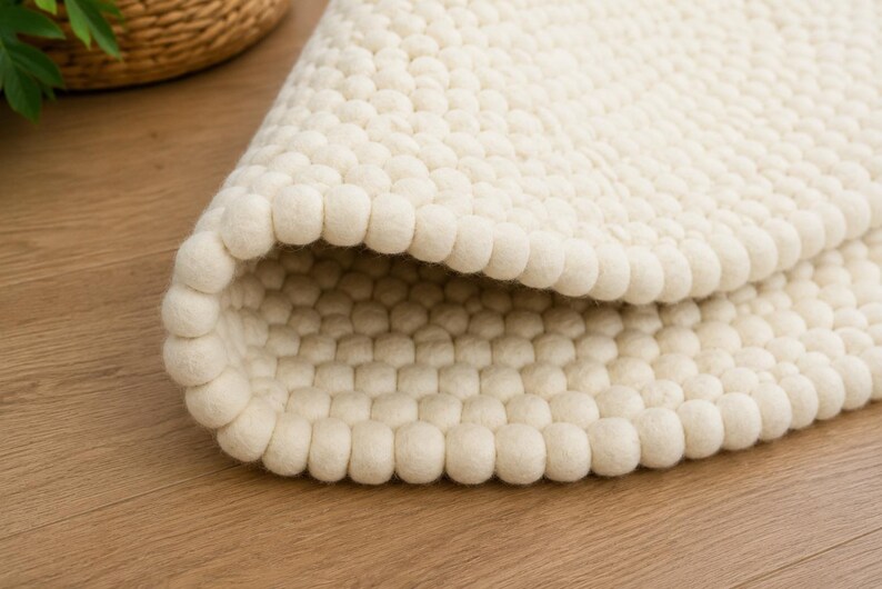 K&ouml;nnte beinhalten: Ein cremefarbener Teppich, der aus zahlreichen kleinen, kugelf&ouml;rmigen Filzwollkugeln in einem dichten, sich wiederholenden Muster besteht. Der Teppich ist teilweise umgeschlagen und zeigt die gleichm&auml;&szlig;ige Textur der Filzkugeln auf beiden Seiten.