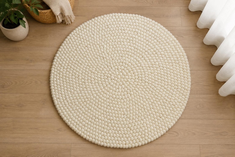 K&ouml;nnte beinhalten: Ein runder Teppich, der aus zahlreichen kleinen, cremewei&szlig;en Filzwollkugeln in einem dichten, konzentrischen Muster besteht. Der Teppich weist eine gleichm&auml;&szlig;ige, texturierte Oberfl&auml;che auf, die durch die einzelnen kugelf&ouml;rmigen Elemente entsteht.