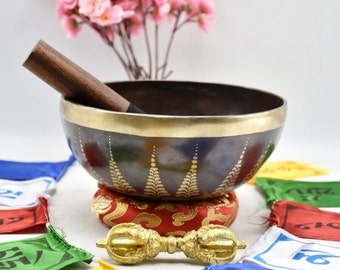 May include: A dark blue singing bowl with a gold rim and intricate gold designs. The bowl is sitting on a red cushion with gold trim. A wooden striker and a gold metal mallet are also included.