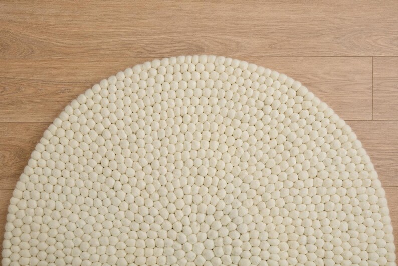 K&ouml;nnte beinhalten: Ein halbkreisf&ouml;rmiger Teppich, der aus zahlreichen kleinen, cremewei&szlig;en Filzwollkugeln in einem dichten, gleichm&auml;&szlig;igen Muster besteht. Die einzelnen Kugeln bilden eine strukturierte, haptische Oberfl&auml;che, die die gesamte runde Form des Teppichs bedeckt.