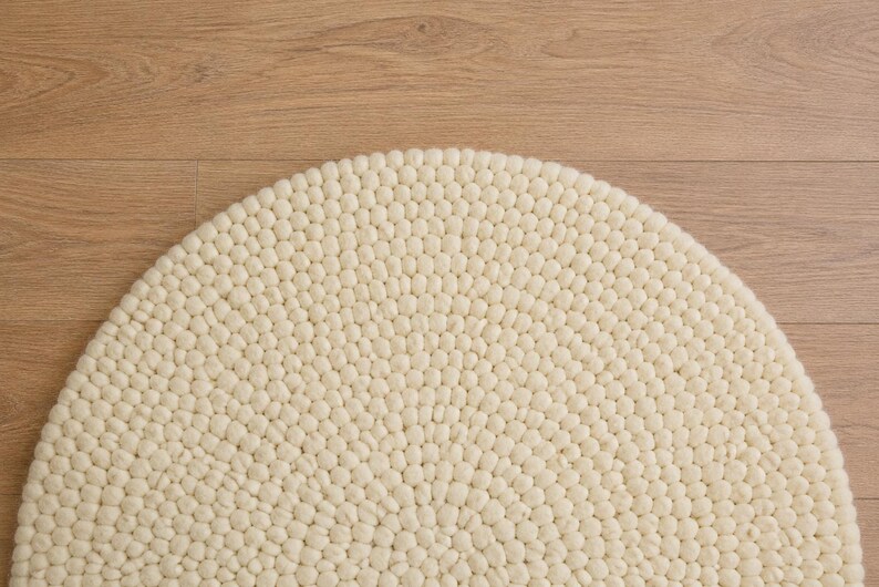 K&ouml;nnte beinhalten: Ein halbkreisf&ouml;rmiger Teppich aus zahlreichen kleinen, cremewei&szlig;en Filzwollkugeln, die in einem dichten, gleichm&auml;&szlig;igen Muster angeordnet sind. Die einzelnen Kugeln bilden eine strukturierte, haptische Oberfl&auml;che auf dem gesamten Teppich.