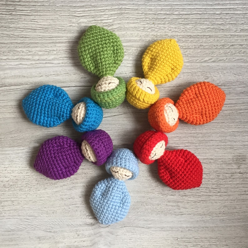 May include: A set of seven crocheted dolls in a rainbow of colors, each with a closed-eye face. The dolls are arranged in a circle on a light wood surface.