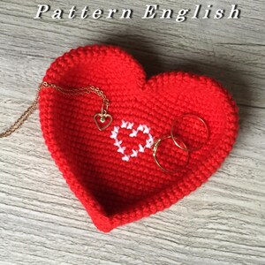 Puede incluir: Plato de corazón rojo tejido a crochet con un diseño de corazón blanco en el centro. El plato está lleno de anillos dorados y un collar dorado con un colgante de corazón.