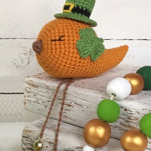 Może przedstawiać: Dziany pomarańczowy ptak w zielonym kapeluszu leprechauna z złotą klamrą i zieloną koniczyną na piersi. Ptak siedzi na stosie białych drewnianych klocków z drewnianym naszyjnikiem z koralików w tle.