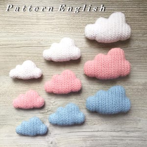 Peut inclure: Une collection de huit nuages en crochet de différentes couleurs, dont le blanc, le rose et le bleu. Les nuages sont disposés en rangée sur une surface en bois.