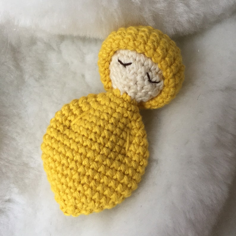 May include: A crocheted doll with a yellow hooded outfit and closed eyes. The doll is lying on a white fluffy surface.